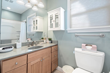 Bright, spa-like modern bathroom with light-blue walls, wood vanity and gray stone countertop with chrome faucet, mirrored medicine cabinet, white shower curtain, wall grab bar and toilet with tissue box.