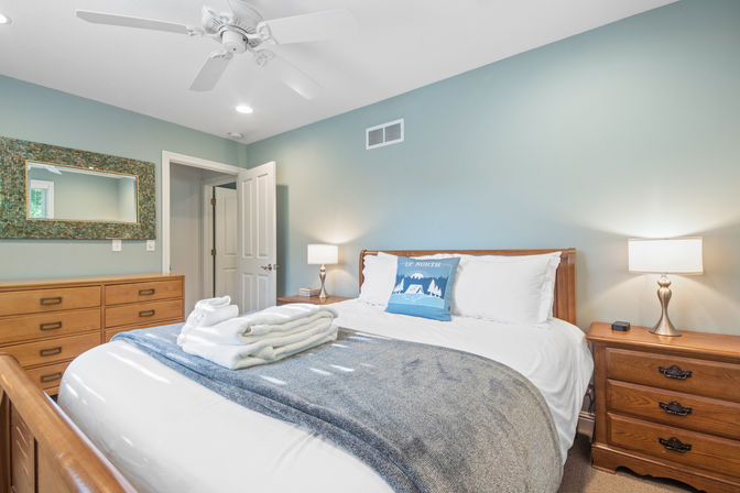 Bright guest bedroom with wooden bed, matching dresser and nightstands; folded towels on bed, blue-gray throw and 'Up North' accent pillow, teal walls, mosaic-framed mirror, ceiling fan and bedside lamps