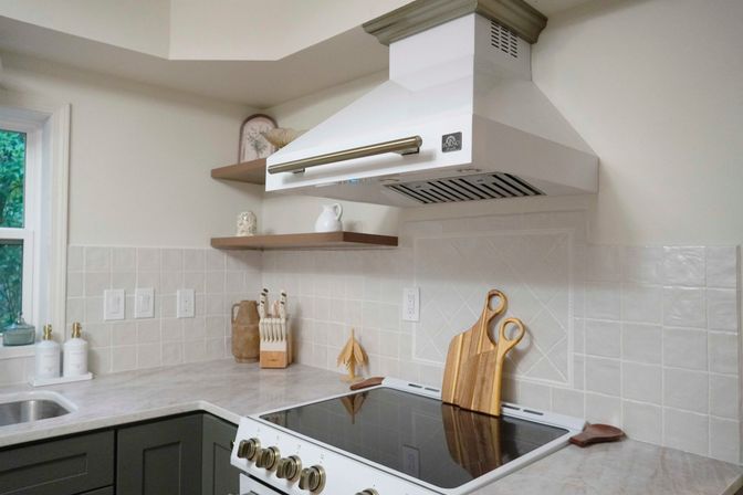 Bright modern farmhouse kitchen corner with white range hood over a black glass cooktop, cream ceramic tile backsplash, floating wood shelves, marble-look countertop, gray lower cabinets and wooden cutting boards.