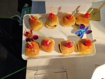 White platter of eight bite-sized mini sliders topped with cherry tomatoes on wooden picks, garnished with purple and magenta orchids — party appetizer platter.