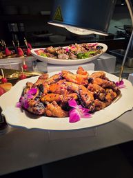 Buffet platter of glazed grilled chicken wings with char marks, garnished with purple orchid flowers on a white serving dish under a heat lamp at a catered food station.