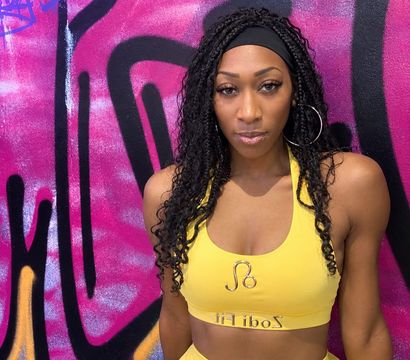 Woman in yellow workout set with braided hair and hoop earrings posing against a vibrant pink and black graffiti mural — urban fitness portrait.