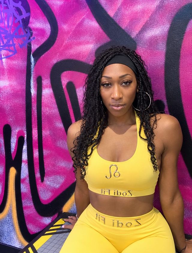 Woman in yellow workout set with braided hair and hoop earrings posing against a vibrant pink and black graffiti mural — urban fitness portrait.