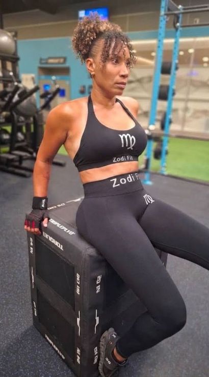 Woman in black sports bra and leggings sits on a plyo box in an indoor gym, wearing workout gloves and sneakers and pausing between strength-training exercises.