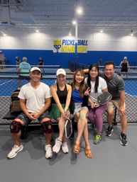 Five smiling adults seated on a bench at an indoor pickleball facility, holding paddles and wearing athletic gear, with blue-walled courts and players in the background.