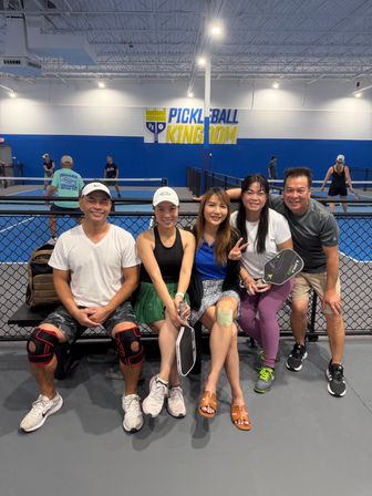 Five smiling adults seated on a bench at an indoor pickleball facility, holding paddles and wearing athletic gear, with blue-walled courts and players in the background.
