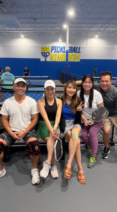 Five smiling adults seated on a bench at an indoor pickleball facility, holding paddles and wearing athletic gear, with blue-walled courts and players in the background.