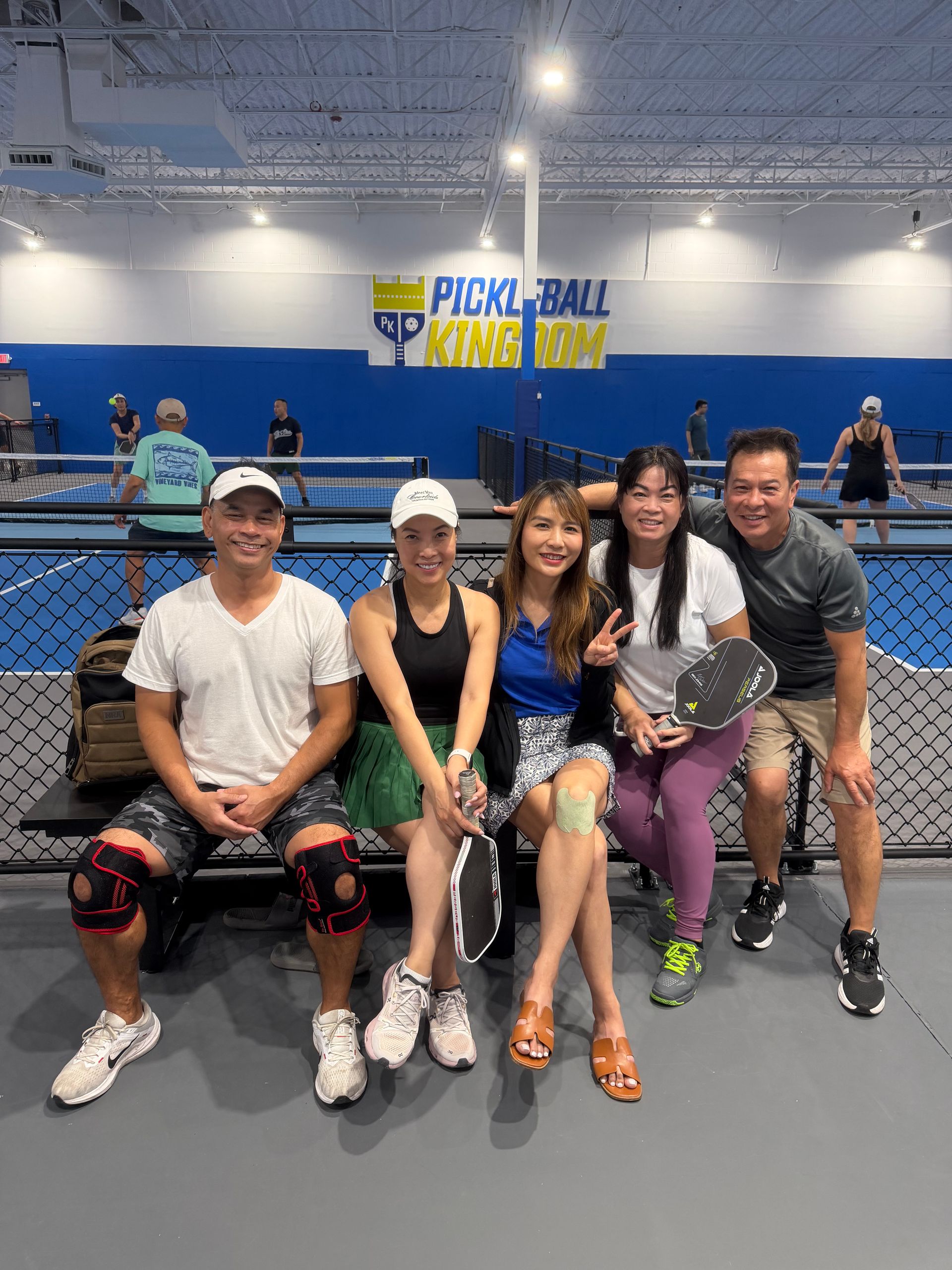 Five smiling adults seated on a bench at an indoor pickleball facility, holding paddles and wearing athletic gear, with blue-walled courts and players in the background.