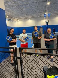 Four players posing with blue-and-yellow pickleball paddles on an indoor pickleball court with bright blue walls and courts 13–14 visible.