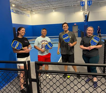 Four players posing with blue-and-yellow pickleball paddles on an indoor pickleball court with bright blue walls and courts 13–14 visible.