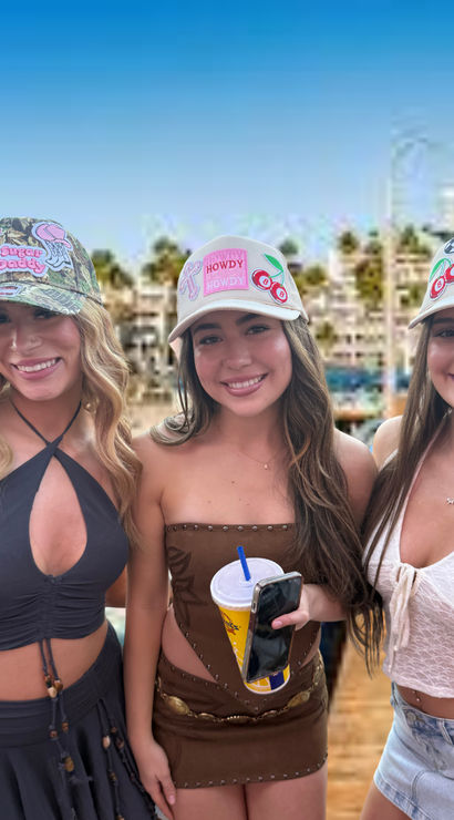 Three smiling young women wearing decorated baseball caps and summer outfits on a sunny marina boardwalk, holding a drink and phone with a blurred palm-lined waterfront and blue sky behind them.