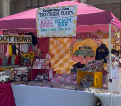 Bright pink pop-up market booth at an outdoor craft fair selling custom trucker hats, boot bows, denim and bandana accessories with sunflower accents and a vintage truck logo display