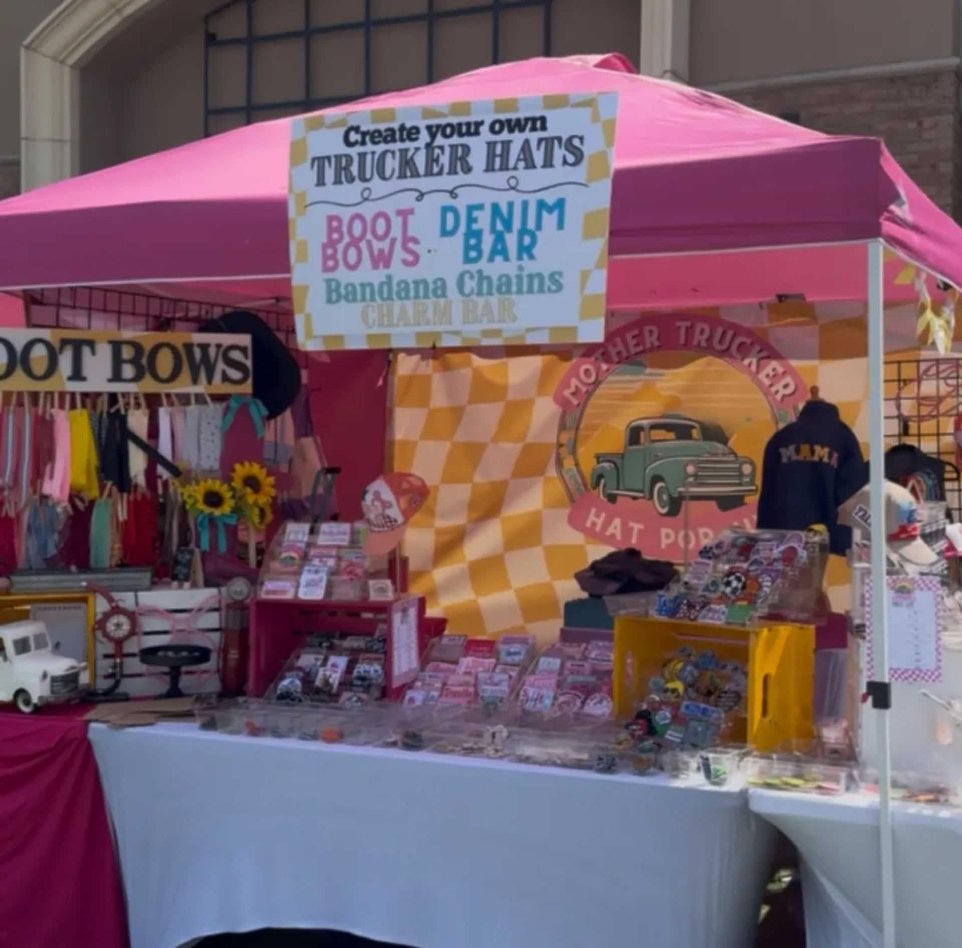 Bright pink pop-up market booth at an outdoor craft fair selling custom trucker hats, boot bows, denim and bandana accessories with sunflower accents and a vintage truck logo display