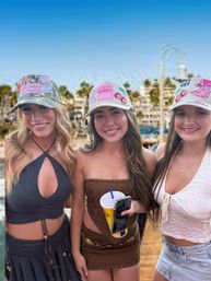 Three smiling young women in colorful trucker hats and summer outfits on a sunny coastal boardwalk, middle holding a drink cup and smartphone with blurred palm trees and beachside buildings behind them.