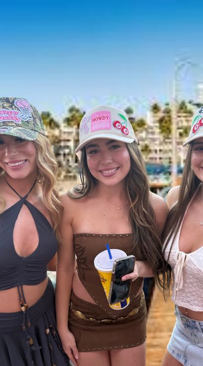 Three smiling young women in decorated trucker hats and summer outfits on a sunny beachfront boardwalk with palm trees, center woman holding a drink and phone.