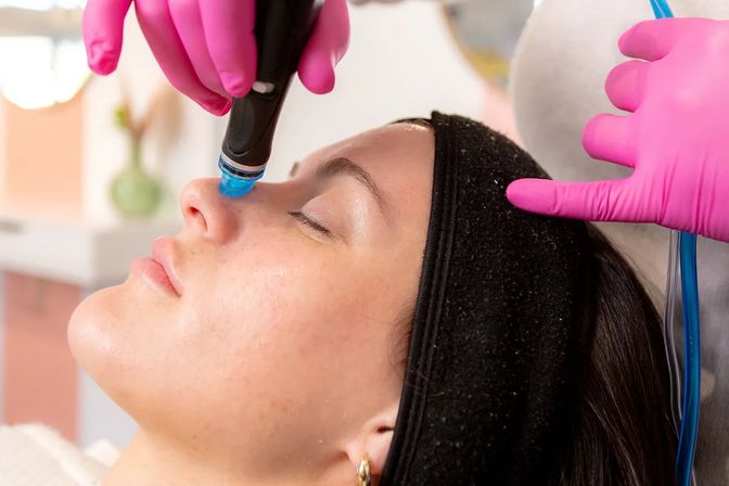 Close-up of a woman receiving a hydrafacial-style facial at a spa, technician in bright pink gloves using a blue-tipped handheld device on her nose.