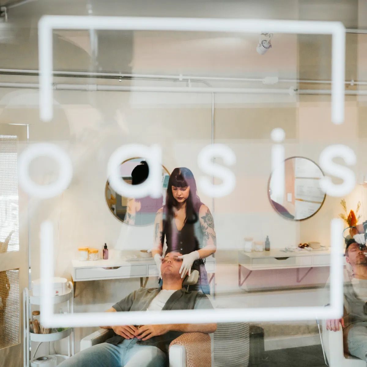 Through a glass window with white logo, a tattooed esthetician in gloves gives a relaxing facial to a reclining client in a boutique spa treatment room with round mirrors and skincare products