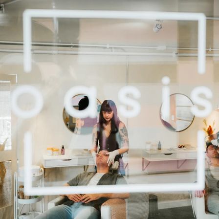 Through a glass window with white logo, a tattooed esthetician in gloves gives a relaxing facial to a reclining client in a boutique spa treatment room with round mirrors and skincare products