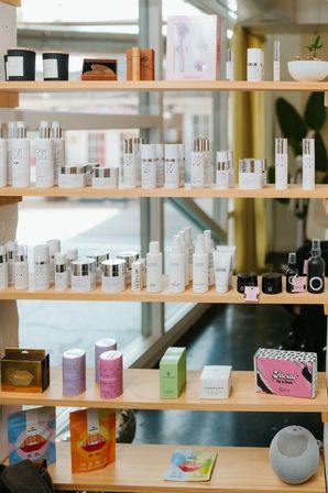 Sunlit boutique shelf of skincare products: rows of white bottles and silver‑lid jars, colorful boxes and face masks arranged on wooden shelves by a large window.