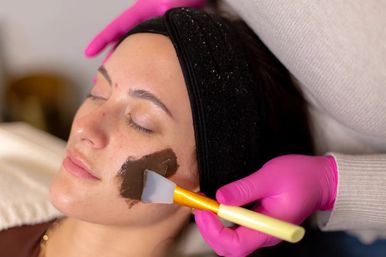 Spa facial treatment: client with black headband relaxes as an esthetician in pink gloves applies a dark clay mask to her cheek with a silicone spatula.