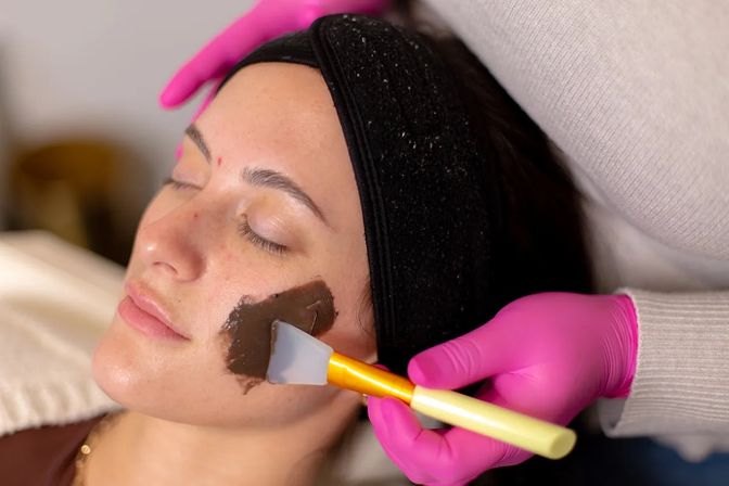 Spa facial treatment: client with black headband relaxes as an esthetician in pink gloves applies a dark clay mask to her cheek with a silicone spatula.