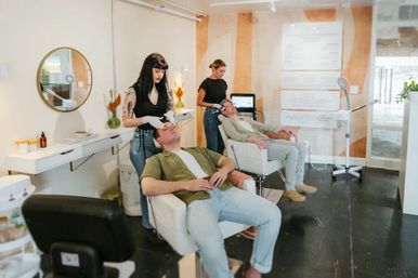 Two men reclining in white salon chairs receiving facial treatments from aestheticians in a bright, modern boutique skincare studio with warm wood accents and treatment equipment.