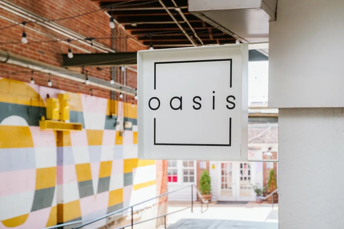 White square hanging sign with a minimalist lowercase logo framed by thin black lines, suspended in a brick-ceiling urban courtyard above a colorful yellow, pink and gray geometric mural and shop entrance.