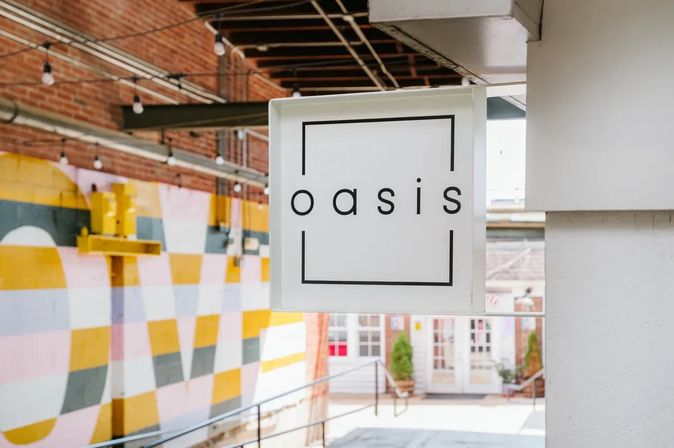 White square hanging sign with a minimalist lowercase logo framed by thin black lines, suspended in a brick-ceiling urban courtyard above a colorful yellow, pink and gray geometric mural and shop entrance.