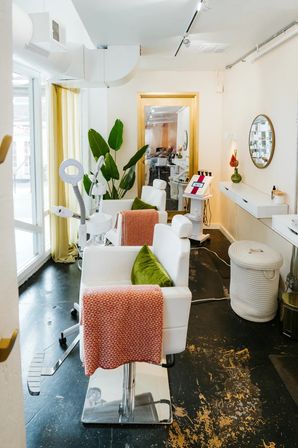 Bright modern beauty salon interior with white styling chairs draped in coral throws and green velvet pillows, magnifying lamp and equipment cart, potted tropical plant, round wall mirror and large front windows letting in natural light.