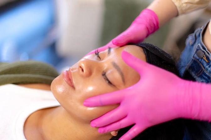 Spa client with dewy skin receiving a relaxing facial massage from an esthetician wearing bright pink gloves.