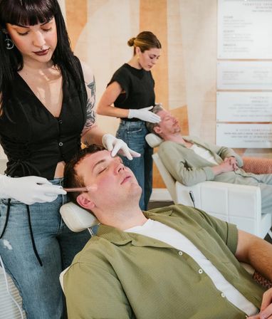 Two men recline in salon chairs while gloved estheticians perform facial treatments with a high-frequency wand in a modern skincare studio