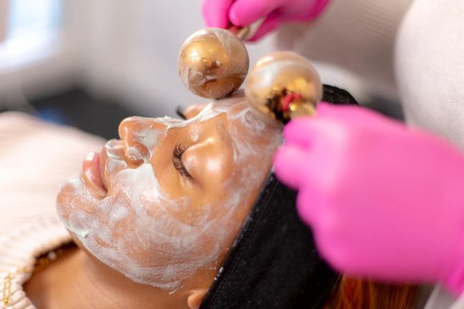 Close-up of a person receiving a spa facial: creamy mask on skin while a technician in bright pink gloves rolls gold globe facial tools across the forehead.