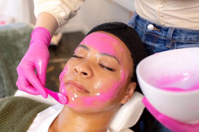 Close-up of a client receiving a bright pink peel-off facial mask at a spa, an esthetician in pink gloves applying glossy skincare with a spatula from a bowl.