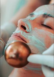 Close-up of a relaxed face with a mint-green facial mask and closed eyes, a gloved hand holding a copper facial roller during a spa skin-care treatment.