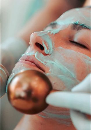 Close-up of a relaxed face with a mint-green facial mask and closed eyes, a gloved hand holding a copper facial roller during a spa skin-care treatment.