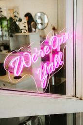 Pink neon 'We're open y'all' sign on a shop window, glowing over a blurred boutique interior with plants and a person in the background