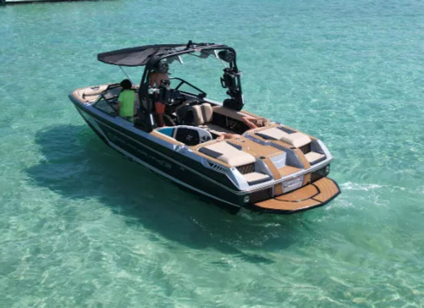 Sleek wakeboard boat with tower, folding canopy and teak swim platform floating in crystal-clear turquoise water with two passengers onboard.