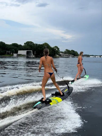 Two people in bikinis riding electric surfboards behind a boat on a lake, cutting twin wakes past a tree-lined shoreline with boathouses under a cloudy sky.