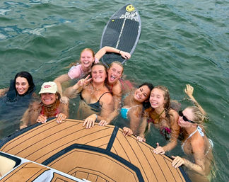 Smiling group of eight young women in swimsuits holding a boat swim platform with a paddleboard nearby on green water — summertime boating and swimming fun.