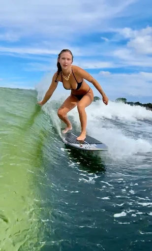 Surfer in a black bikini crouched on a surfboard carving a translucent green ocean wave under a bright blue sky — action surfing on a coastal summer day.