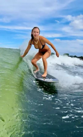 Surfer in a black bikini crouched on a surfboard carving a translucent green ocean wave under a bright blue sky — action surfing on a coastal summer day.
