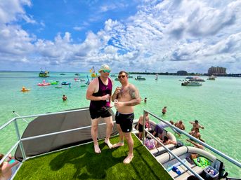 Two friends on the upper deck of a pontoon boat holding drinks, overlooking a crowded turquoise bay with boats, colorful inflatable floats and swimmers under a sunny, partly cloudy sky — lively coastal boat party scene.