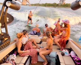 Friends on a wake boat watching and filming a man wakesurfing in the boat’s wake on a calm lake at sunset