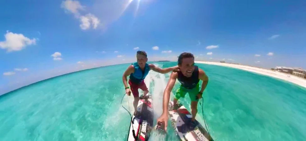 Two friends riding motorized surfboards across clear turquoise tropical water toward a white sandy beach under a bright blue sky, smiling and enjoying watersports.