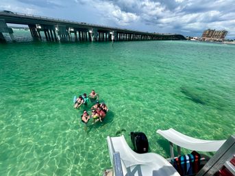 Group of people floating in clear turquoise bay near a boat's white slide, with a long concrete bridge and beachfront buildings under a partly cloudy sky.