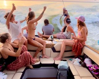 Group of friends cheering on a motorboat as a wakesurfer rides the wake at sunset on a lake — sunlit summer boat party vibes.
