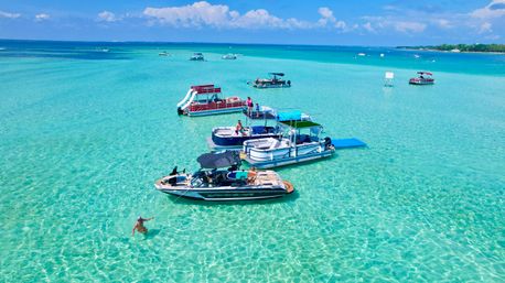 Aerial view of pontoon and motor boats anchored in crystal-clear turquoise shallow water, people relaxing on decks and a swimmer holding a rope under a sunny blue sky
