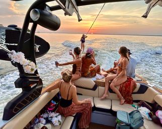 Group of people on a wakeboat's stern at sunset, watching and filming a wakesurfer riding the boat's wake across calm coastal waters with a vibrant orange sky.