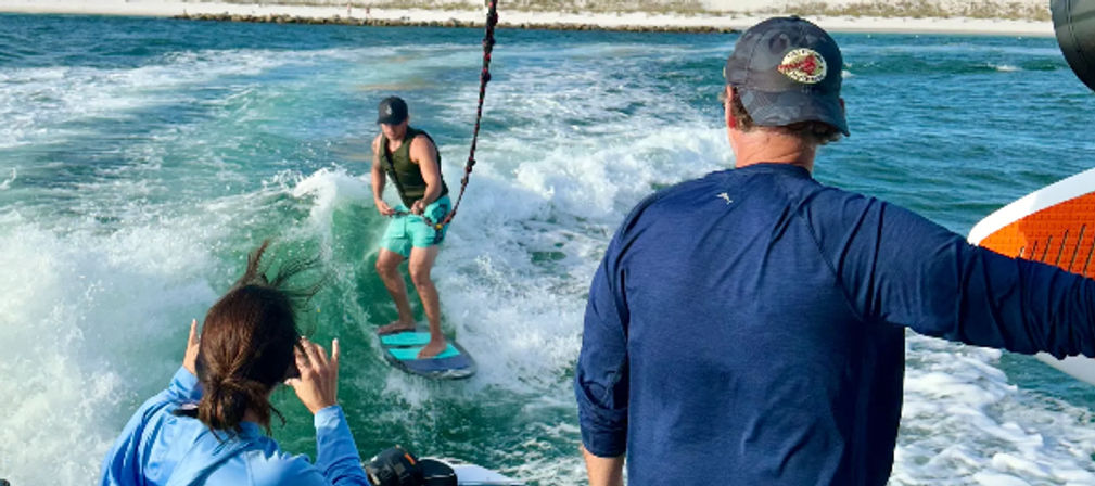Wakesurfer riding a blue board on a boat’s breaking wake in turquoise coastal water, two people on the boat watching and holding a surfboard with a sandy shoreline in the background.