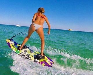 Bikini-clad wakeboarder riding a colorful board across turquoise ocean, kicking up spray with boats on the distant horizon.